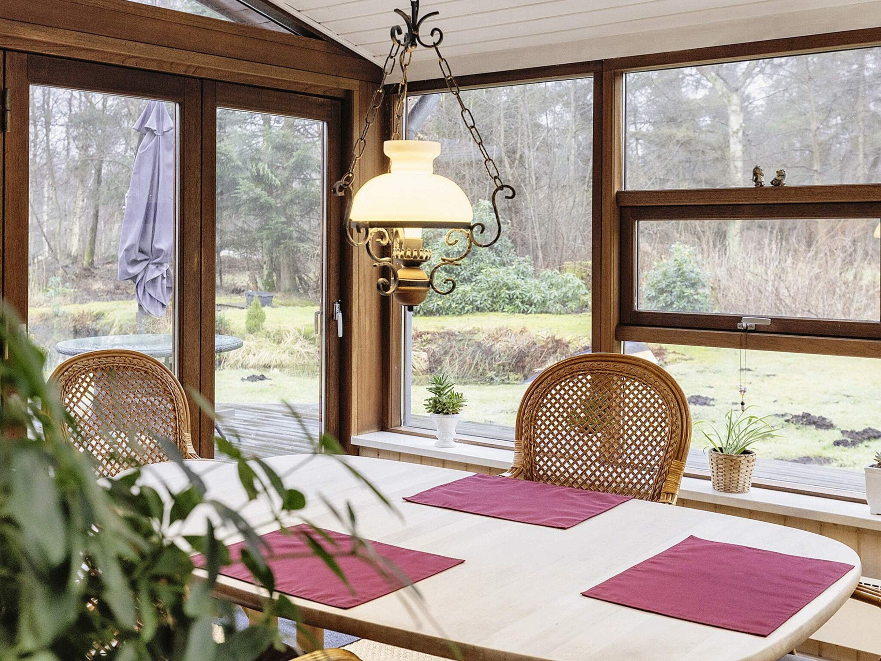 A room with a table set for two, two wicker chairs, a hanging light fixture, and a view of a garden through large windows.