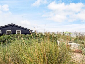 A dark-colored building with a simple, modern design, situated in a natural setting with tall grasses and a body of water in the background. - 4 Star Holiday Home in Karrebaeksminde (Karrebæksminde)