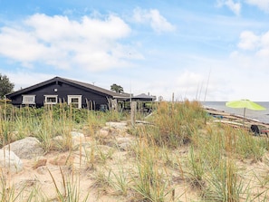 A beach house with a dark exterior, a covered outdoor area, and a sandy foreground with tall grasses. - 4 Star Holiday Home in Karrebaeksminde (Karrebæksminde)