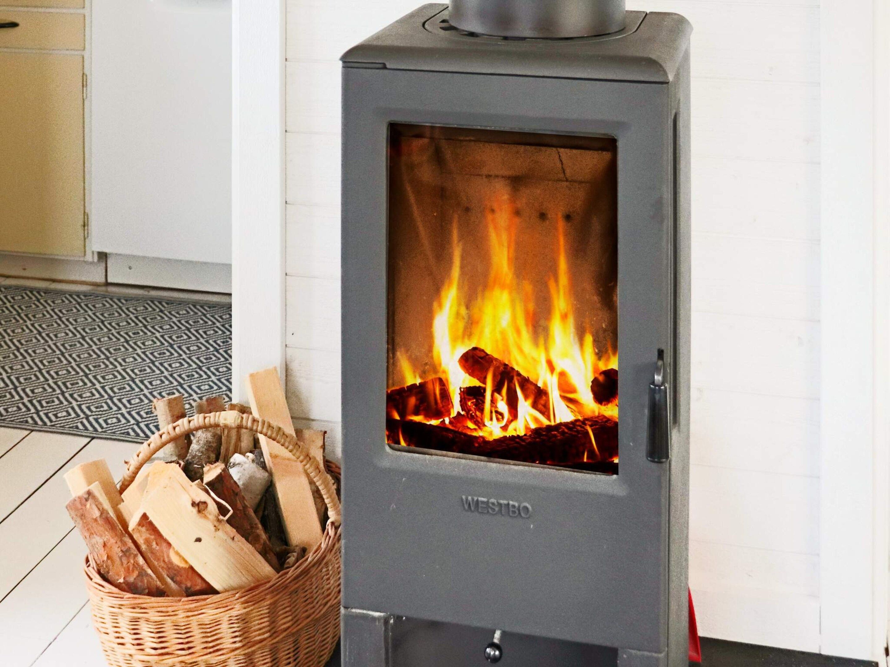 En Westbo-vedeldad kamin med en brasa som sprakar inuti, placerad bredvid en korg av rotting som är fylld med ved.