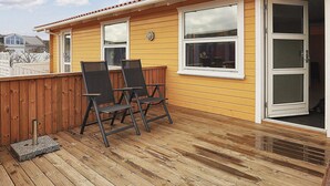 Um deck de madeira com duas cadeiras e uma mesa pequena, uma porta de vidro deslizante e uma janela com vista para o céu.