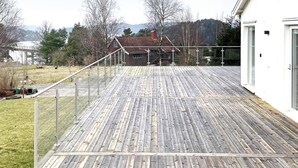 A wooden deck with a metal railing, a white building with glass doors, and a view of a house with a red roof in the background. - 4 Star Holiday Home in Uddevalla (Uddevalla)