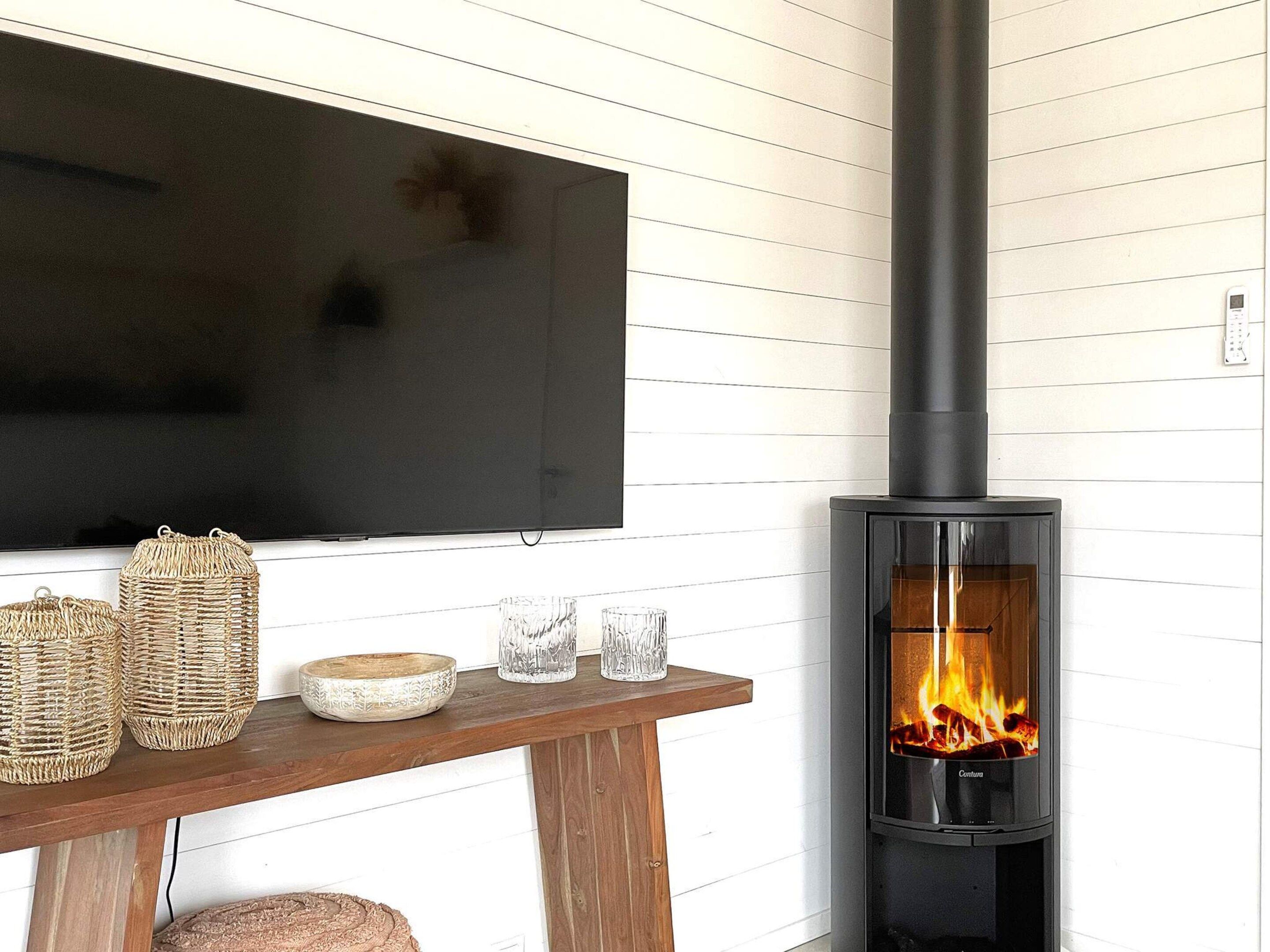 A living room with a flat-screen TV mounted on the wall, a wooden table with decorative items, and a black wood-burning stove with visible flames.