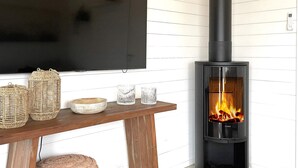 A living room with a flat-screen TV mounted on the wall, a wooden table with decorative items, and a black wood-burning stove with visible flames. - 4 Star Holiday Home in Uddevalla (Uddevalla)