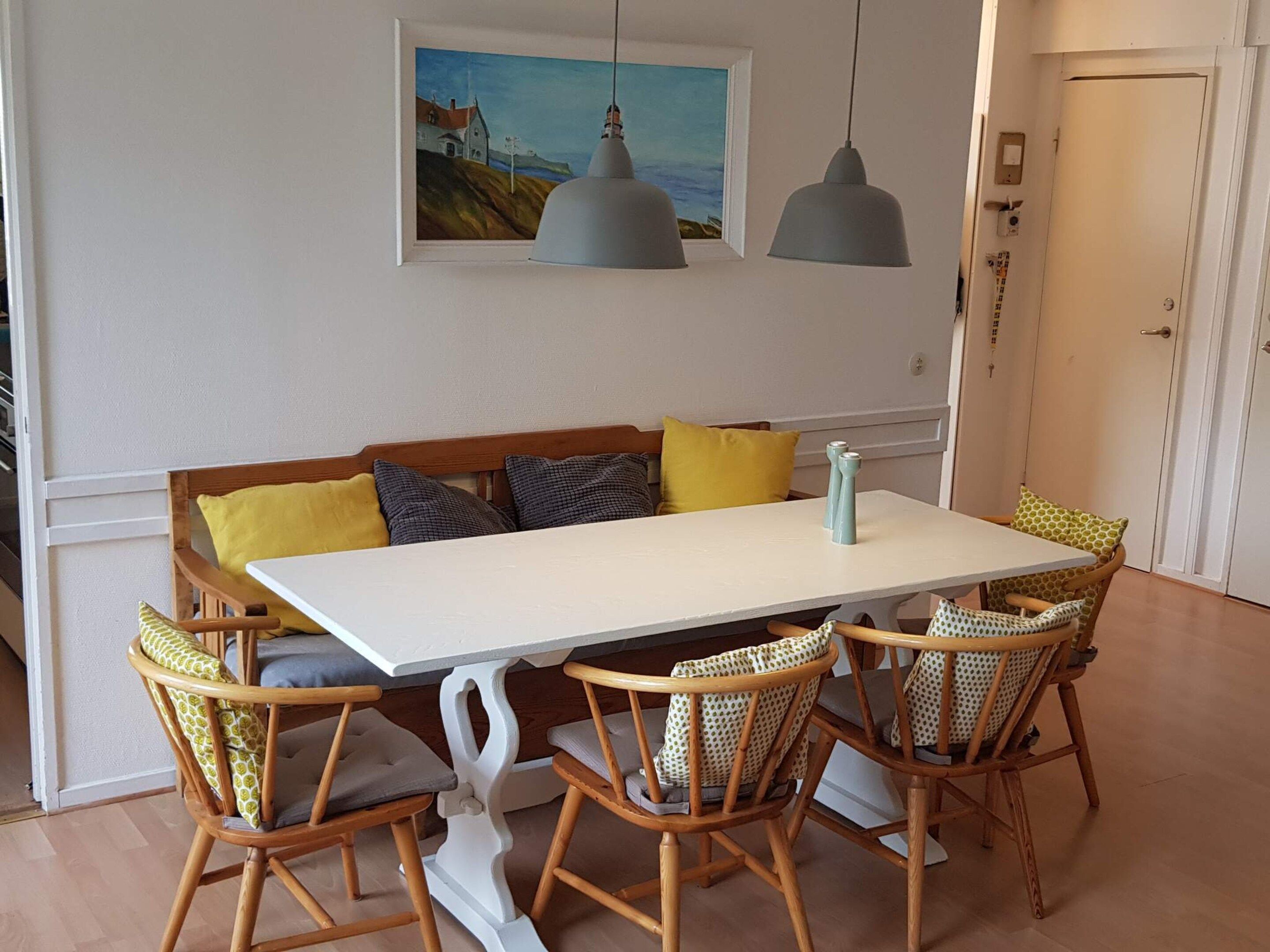 A dining area with a white table, four wicker chairs, a bench with cushions, and two pendant lights.