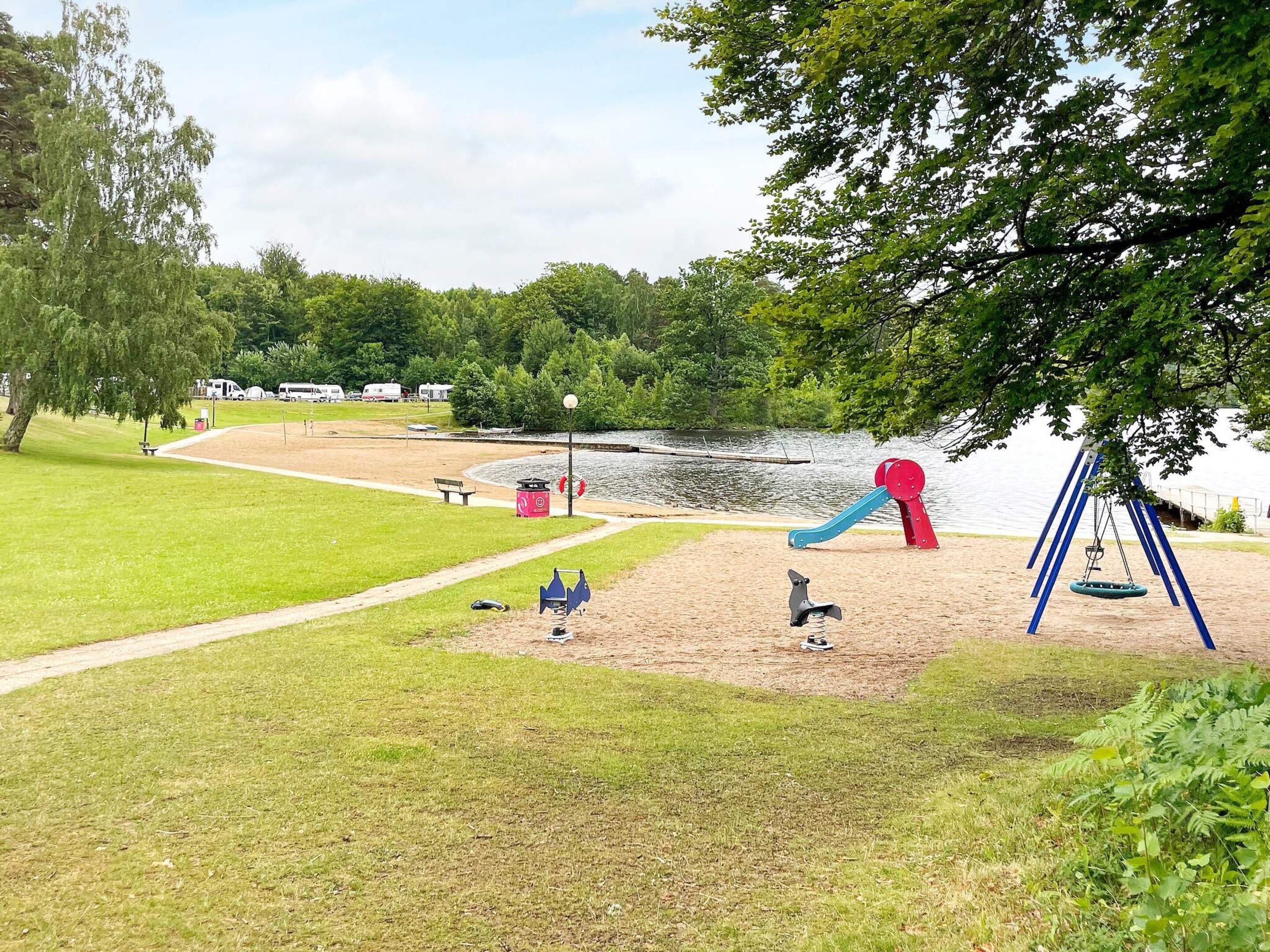 Un parque con un área de juegos, zona arenosa, sendero y un lago al fondo.
