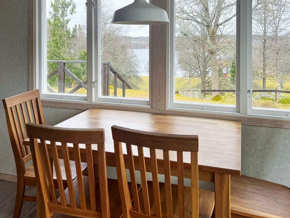 A dining area with a wooden table and chairs, large windows with a view of trees and a lake, and a pendant light.