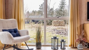A modern living room with a rocking chair, a large window, and a view of a yard with trees and a stack of logs. - 4 Star Holiday Home in Albaek (Aalbaek)