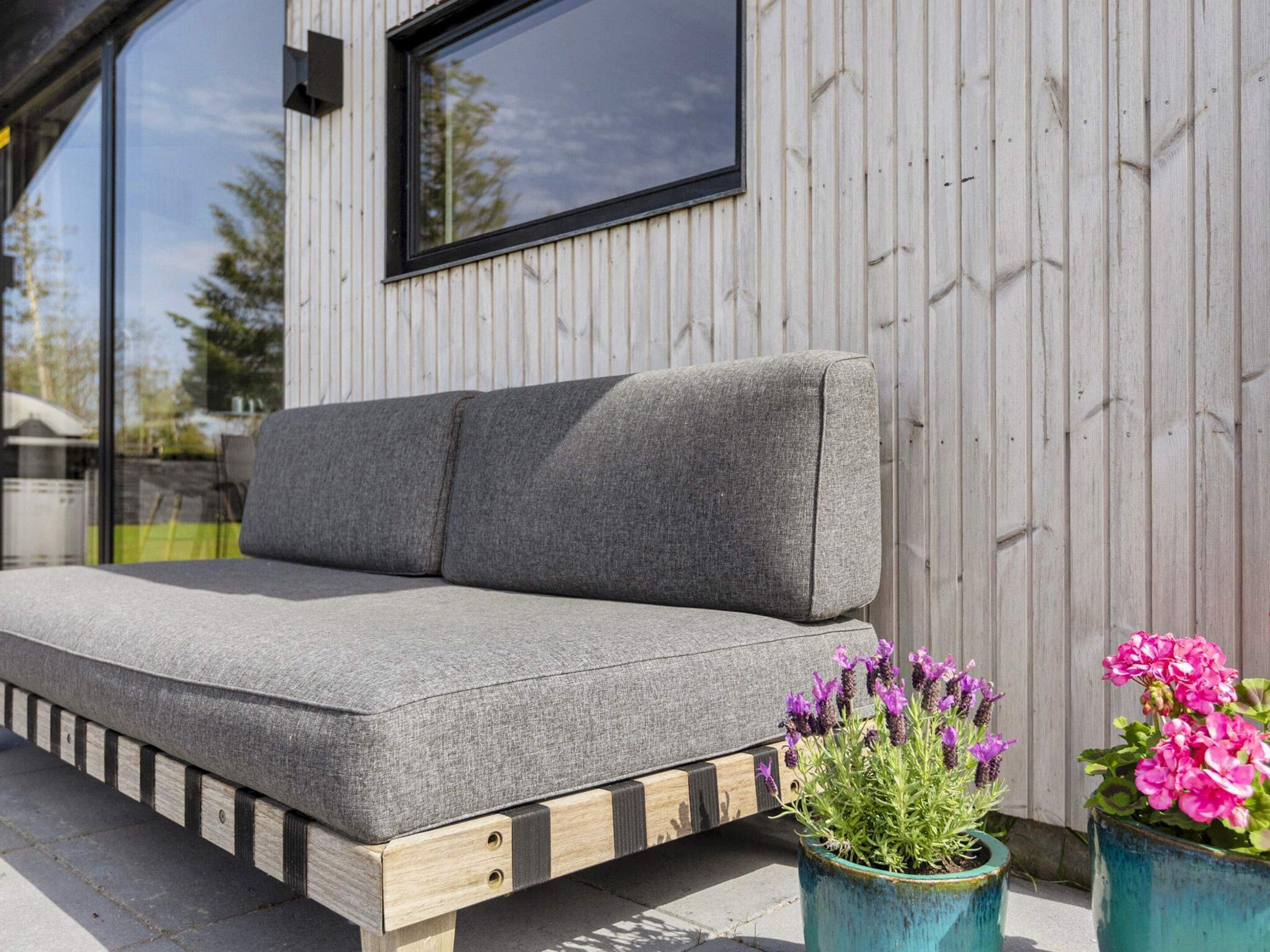 A grey sofa with wooden legs and a cushion, positioned on a concrete surface with two potted plants beside it.