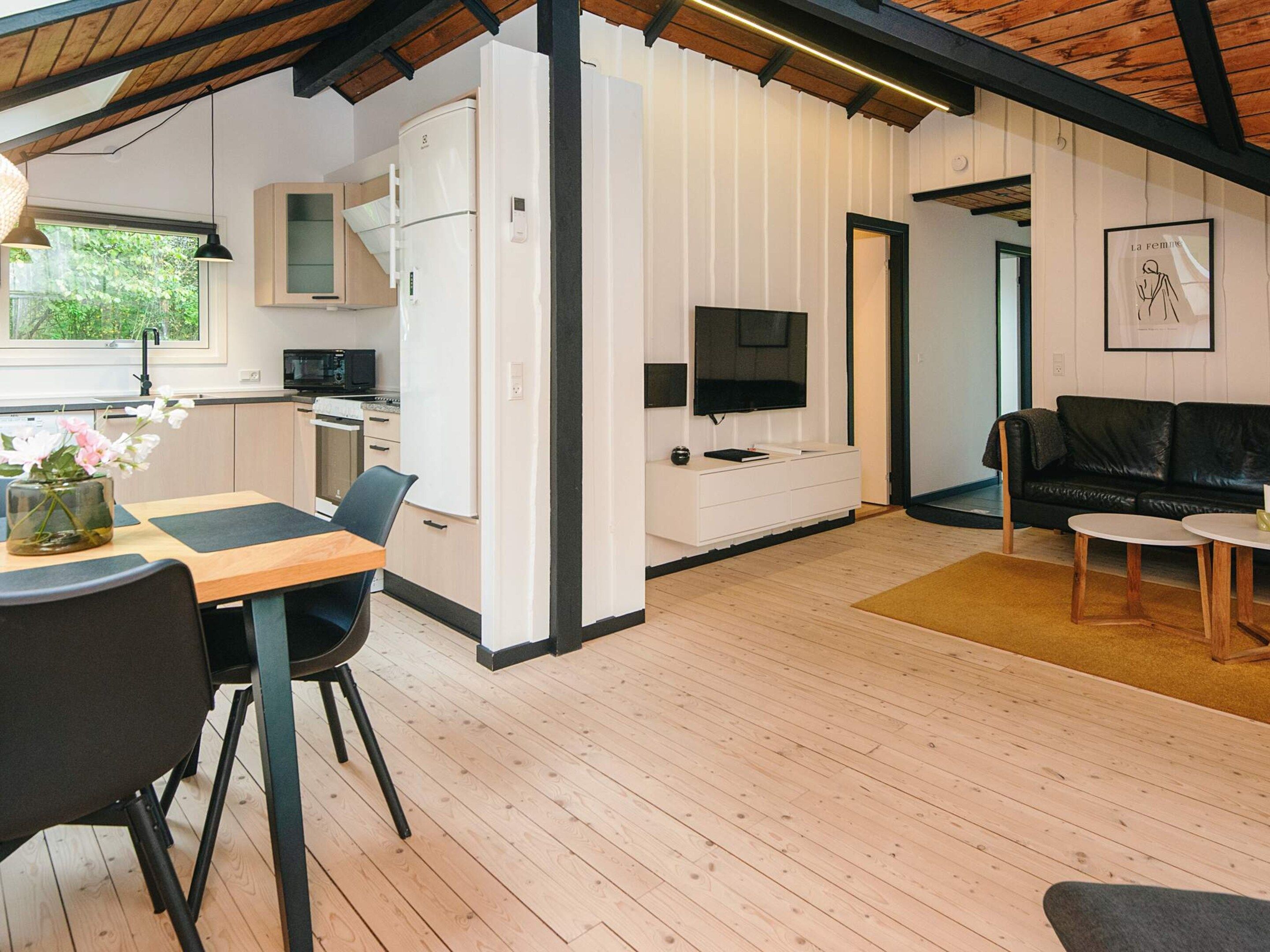 A modern kitchen with white cabinets, a dining area with a table and chairs, a black leather sofa, and a television.