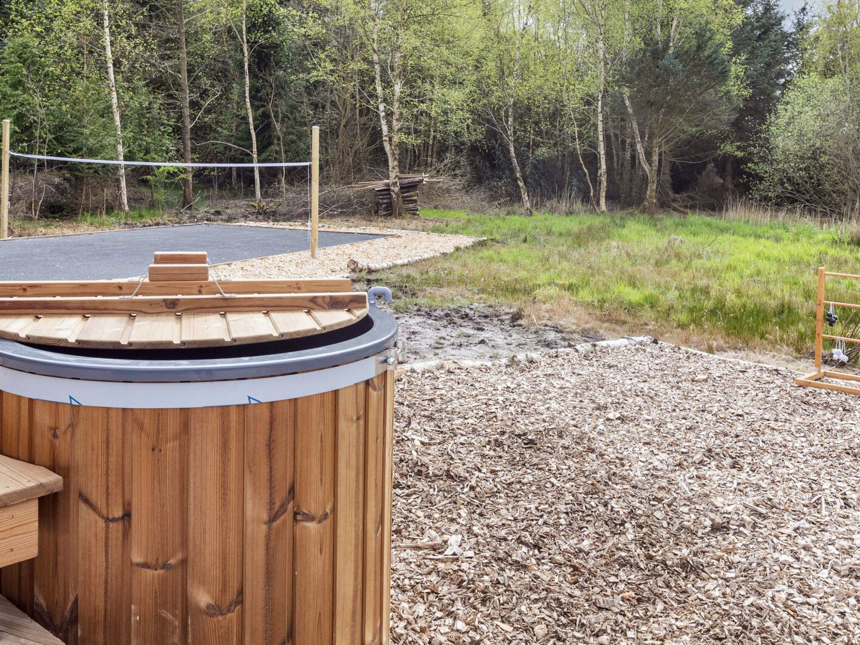 A wooden sauna structure with a sloped roof, surrounded by a gravel area and a forested backdrop.