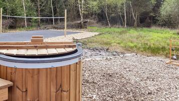 A wooden sauna structure with a sloped roof, surrounded by a gravel area and a forested backdrop.