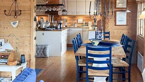 A cozy cabin-style interior with a wooden dining table, blue chairs, and a kitchen area with wooden cabinets and hanging utensils. - 10 Person Holiday Home in Skjaerhalden (Hvaler)