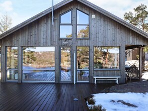 A wooden cabin with large windows, a deck, and a covered outdoor seating area.
