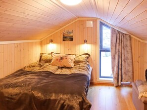 A cozy attic bedroom with a wooden sloped ceiling, a large bed with patterned bedding, two wall-mounted lights, and a window with curtains.