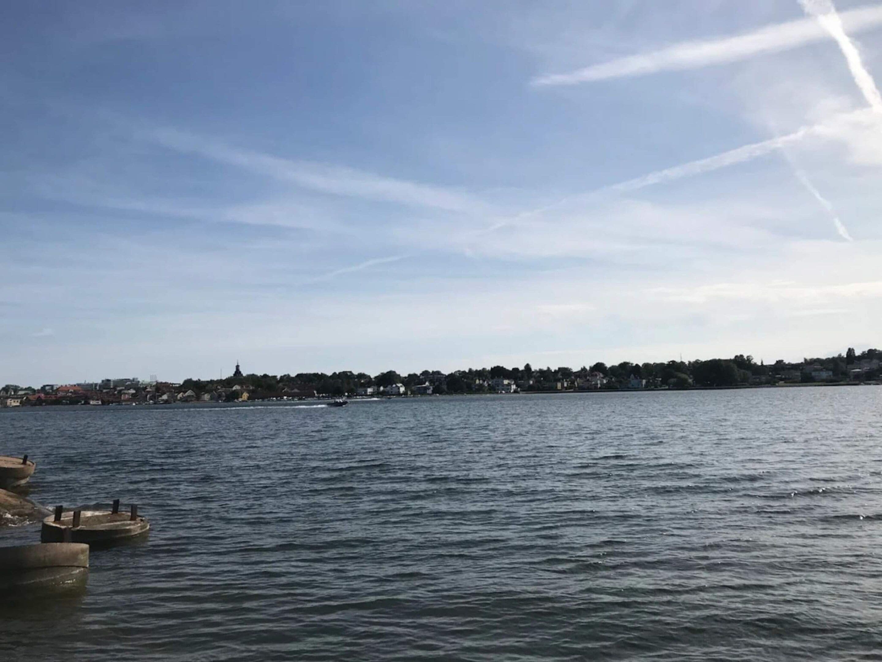 Une vue sur le bord de l’eau, avec une étendue d’eau calme, une côte lointaine parsemée de bâtiments et un ciel dégagé.