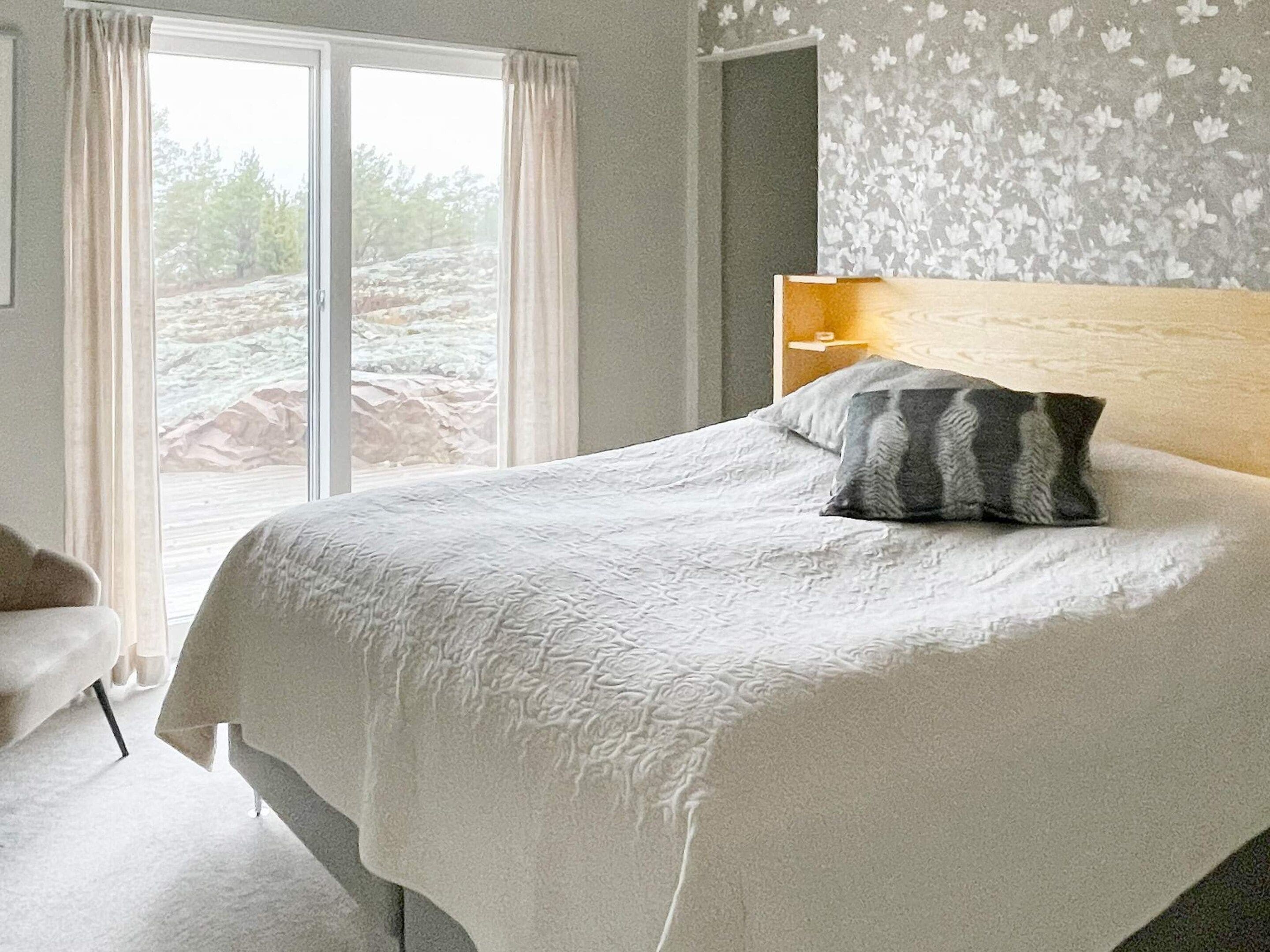 A bedroom with a large bed, a nightstand, a chair, and a view of the outdoors through a window.
