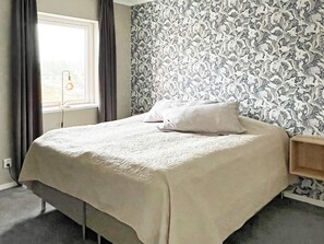 A single bed with a white bedspread and a pillow, a bedside table, a window with curtains, and a patterned wallpaper. - 5 Star Holiday Home in Vastervik (Vastervik)