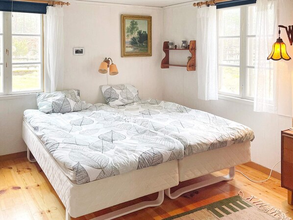 Un dormitorio con cama, dos ventanas, un cuadro, una estantería con adornos y una lámpara.