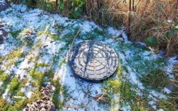 Ett metallgaller på snötäckt mark, omgivet av gräs och buskar.