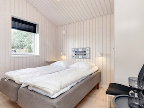 A single bed with a grey mattress and white bedding, a bedside table with a lamp, a wall-mounted power outlet, and a window with blinds. - 4 Star Holiday Home in Vaeggerlose (Vaeggerlose)