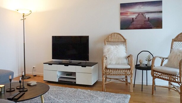 A living room with a flat-screen TV, wicker chairs, a white sofa, a floor lamp, and a painting of a pier.