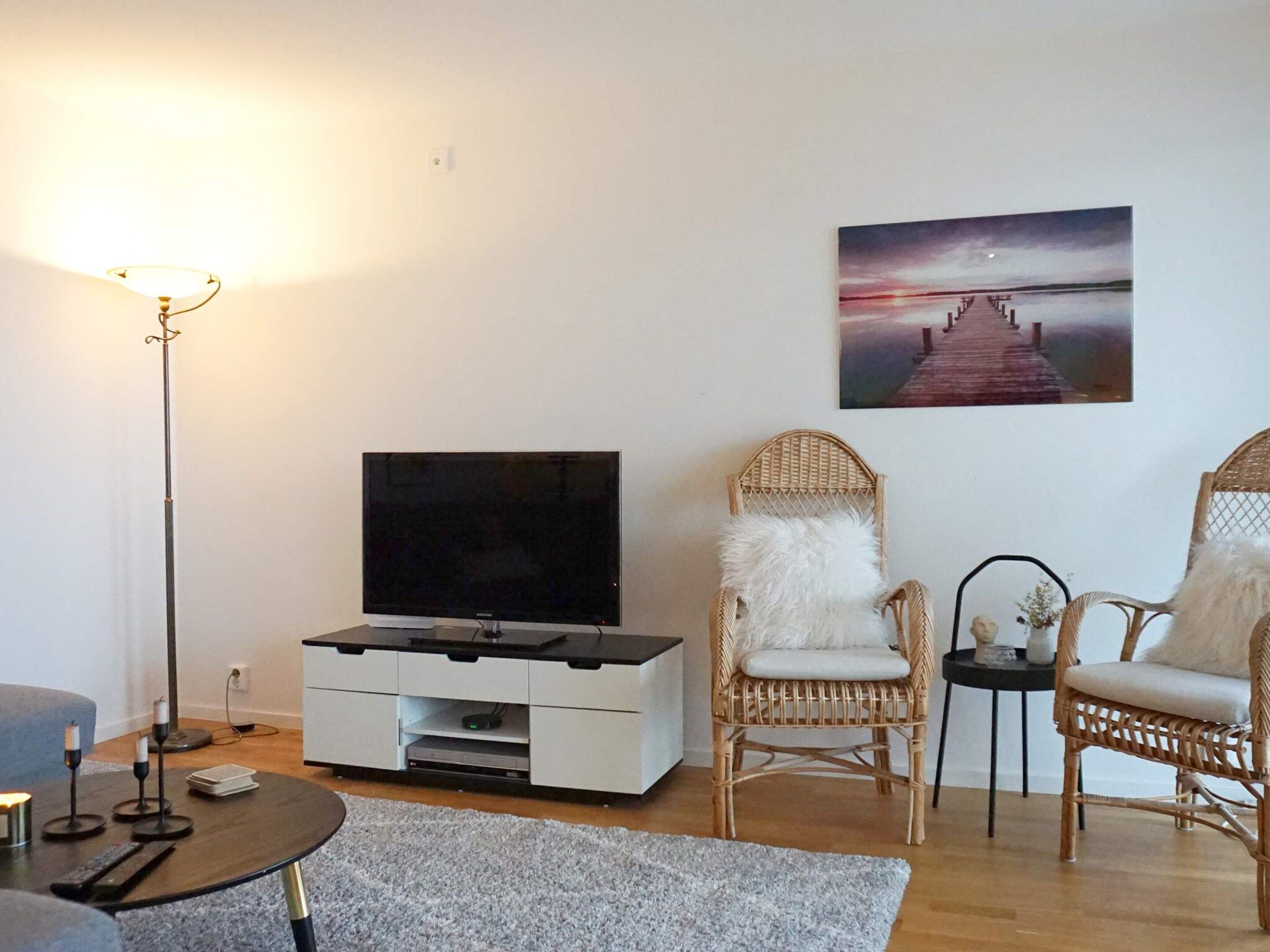 Una sala de estar con un televisor de pantalla plana sobre un mueble blanco, sillas de mimbre, un tapete gris y un cuadro de un muelle en la pared.