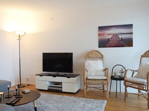 Una sala de estar con un televisor de pantalla plana sobre un mueble blanco, sillas de mimbre, un tapete gris y un cuadro de un muelle en la pared.