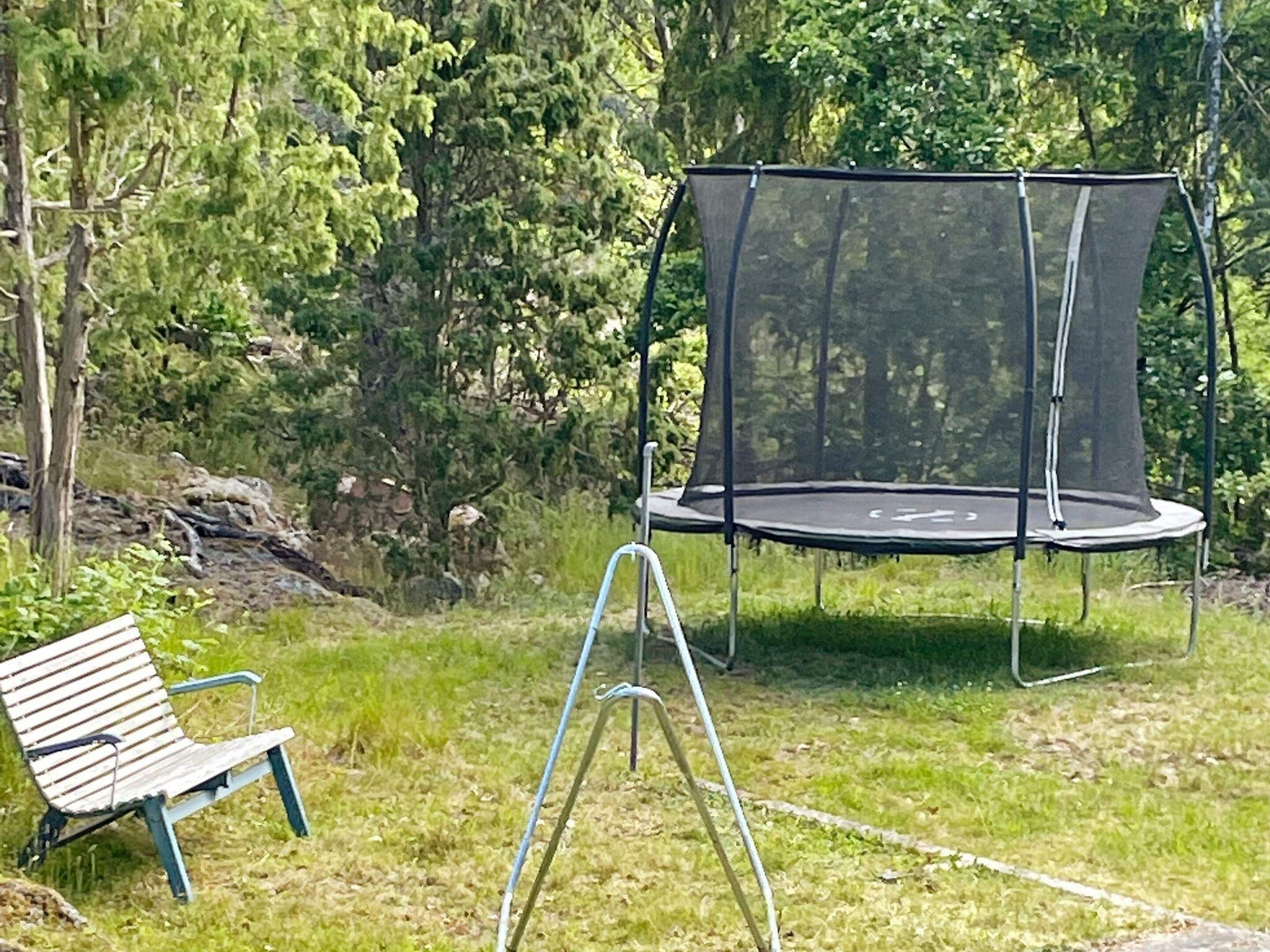 Een trampoline met veiligheidsnet, een bankje en een gazon met bomen op de achtergrond.