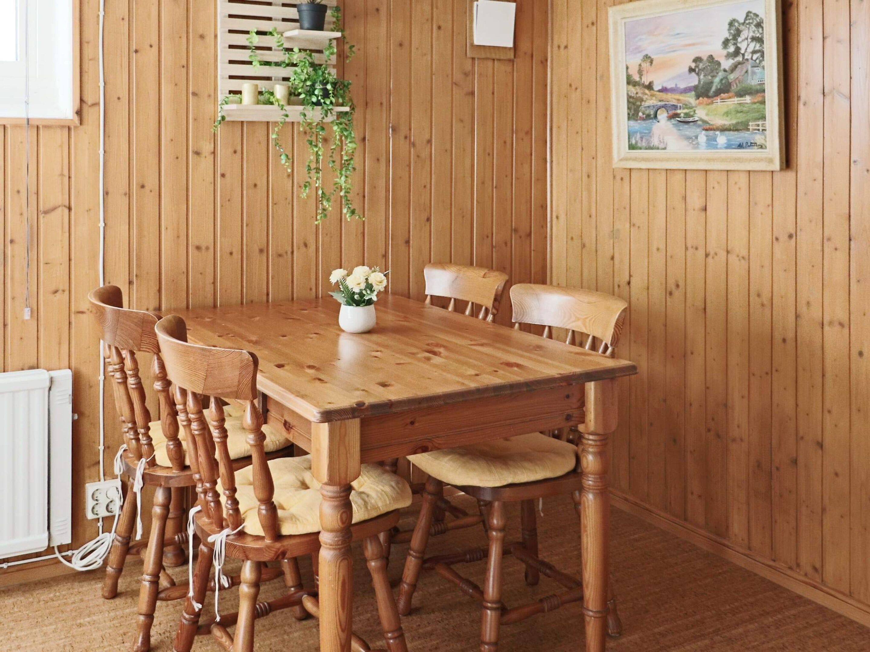 A wooden dining table with six chairs, a white vase with flowers, and a framed landscape painting on the wall.