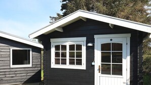 A small, dark wooden cabin with a white door and two windows, surrounded by a grassy area with rocks. - 6 Person Holiday Home in Hadsund (Hadsund)