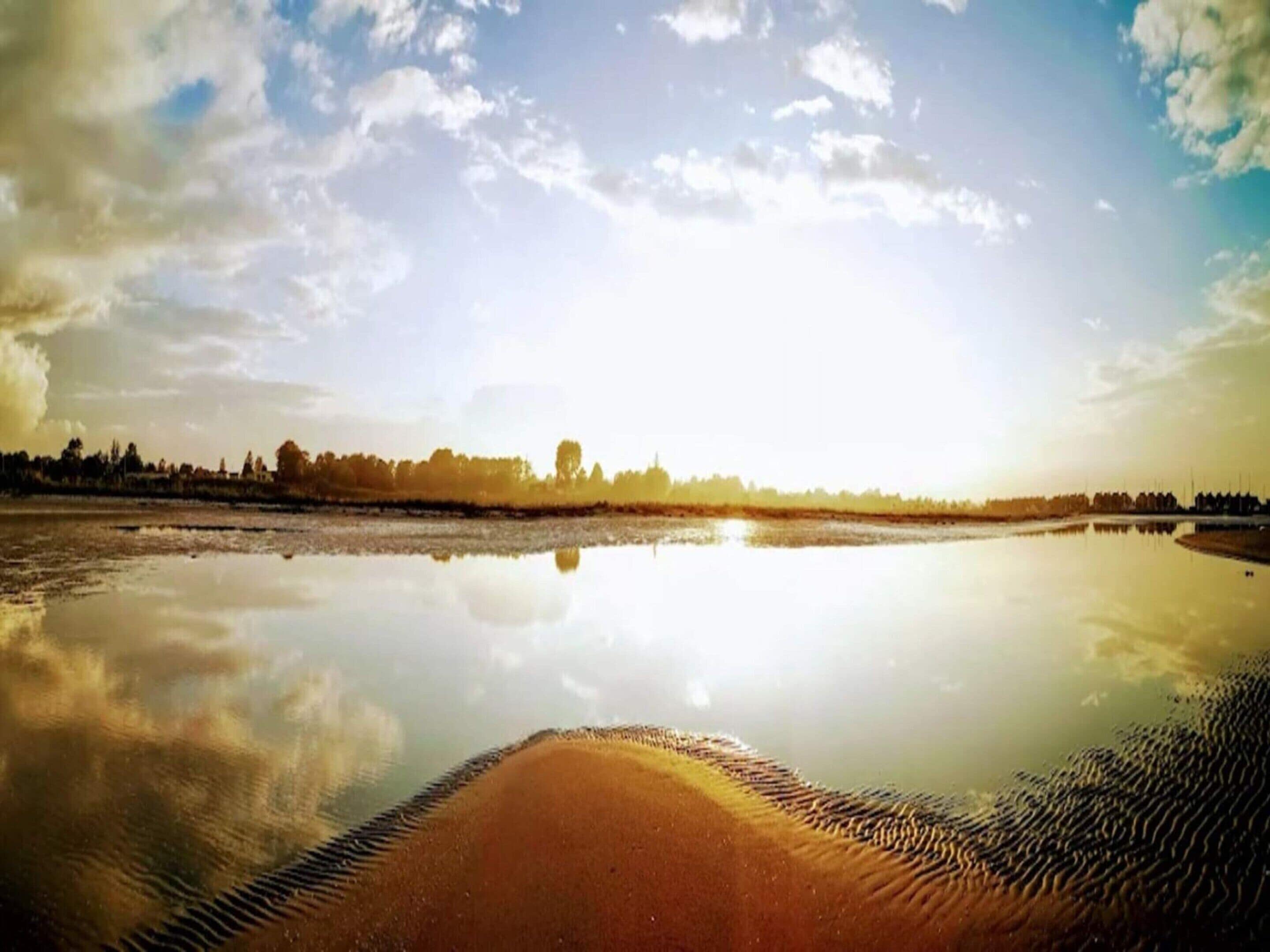 A calm water body reflecting the sky, surrounded by a flat landscape with distant trees and a clear horizon.