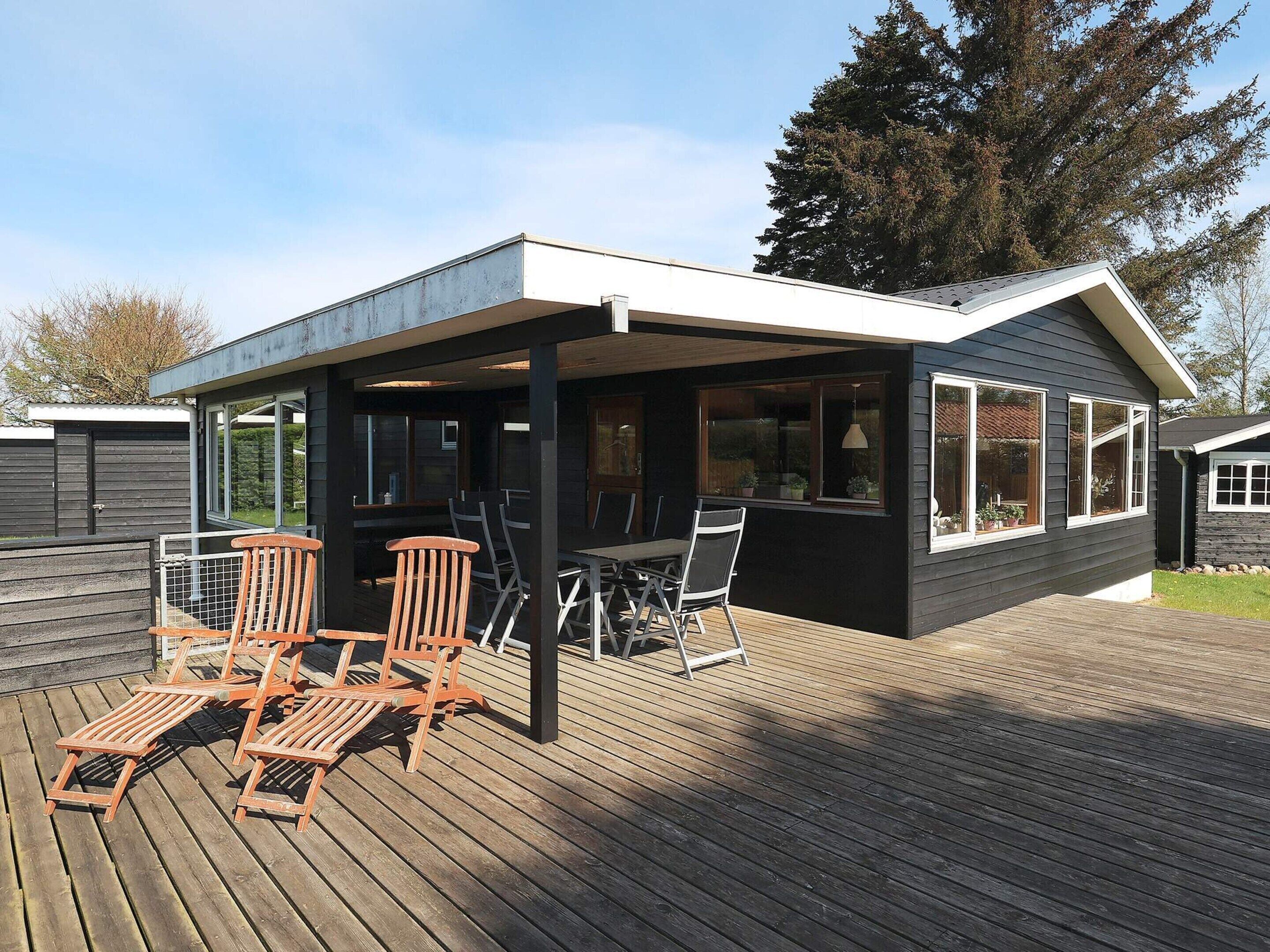 A covered outdoor seating area with wooden chairs and a table, adjacent to a building with large windows.