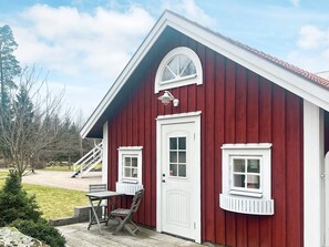 En röd träbyggnad med en vit dörr, två fönster och en liten veranda med ett bord och några stolar.