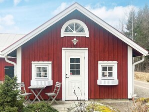 En röd träbyggnad med en vit dörr, två fönster och en liten veranda med ett bord och stolar.