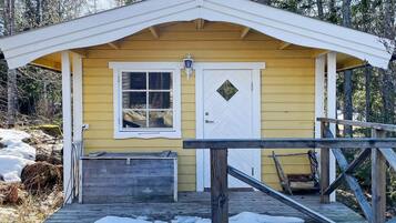 A small yellow cabin with a white door and window, a wooden porch, and a snow-covered deck.