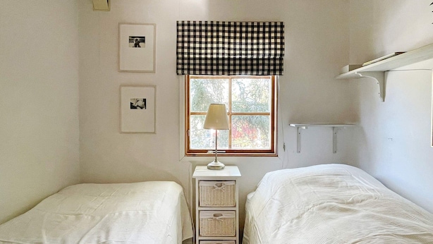 Une chambre avec deux lits simples, une fenêtre agrémentée d’une lambrequin à carreaux, une table de chevet avec une lampe et des étagères fixées au mur.