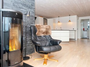 A modern living room with a fireplace, a black leather chair with a fluffy pillow, and a kitchen in the background. - 5 Star Holiday Home in Vinderup (Vinderup)
