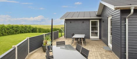 Une terrasse avec une table et des chaises, une porte donnant sur un espace abrité et une vue sur un champ verdoyant.