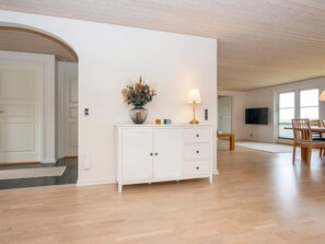 Una habitación amplia con suelo de madera, un armario blanco con cajones, un jarrón con plantas secas, una mesa con sillas y un televisor montado en la pared.