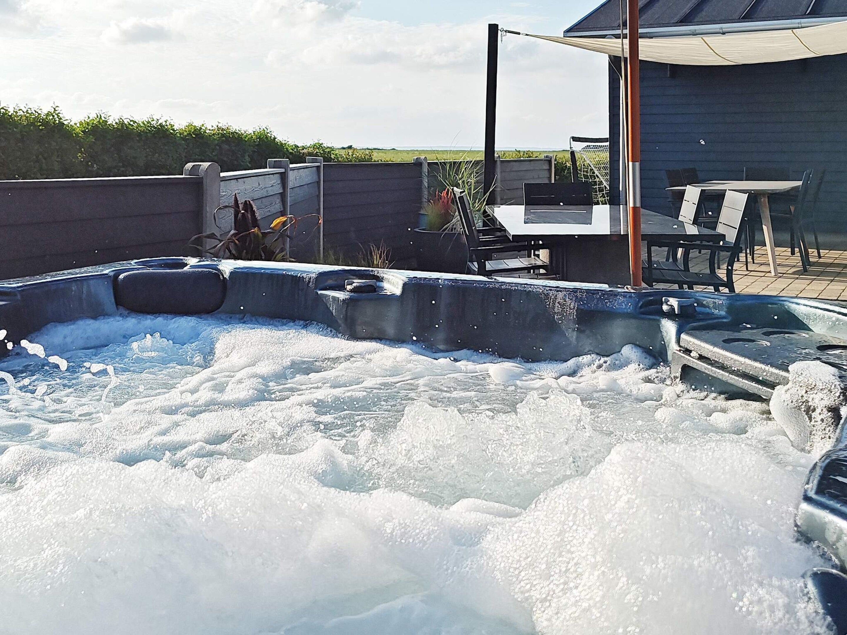 Un jacuzzi rodeado por una terraza de madera, con una parrilla y mobiliario exterior al fondo.