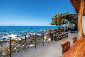 A modern outdoor seating area with a view of the ocean, stone walls, and a wooden table. - Hale Kale:Direct Oceanfront Masterpiece,Modern Spectacular One of a Kind Home (KAILUA KONA)