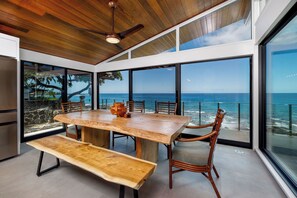 A modern dining area with a large wooden table and chairs, large windows with ocean views, and a ceiling fan. - Hale Kale:Direct Oceanfront Masterpiece,Modern Spectacular One of a Kind Home (KAILUA KONA)