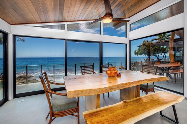 A modern dining area with a large wooden table and chairs, large windows offering a view of the ocean, and a ceiling fan.