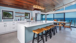 A modern kitchen with a large island, wooden stools, and a view of the ocean. - Hale Kale:Direct Oceanfront Masterpiece,Modern Spectacular One of a Kind Home (KAILUA KONA)