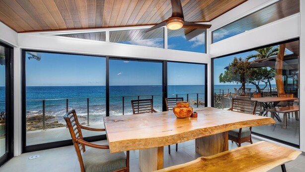 A modern dining area with a large wooden table and chairs, large windows offering a view of the ocean, and a ceiling fan. - Hale Kale:Direct Oceanfront Masterpiece,Modern Spectacular One of a Kind Home (KAILUA KONA)