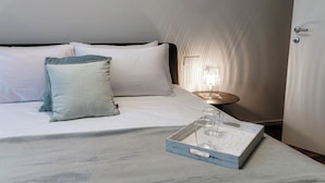 A neatly made bed with white linens and a grey throw, a bedside table with a lamp, and a tray with glasses and a book. - Marinetti Suite 8 (Como)