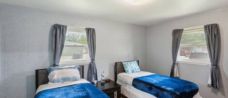 Ein Zimmer mit zwei Betten, jeweils mit einer blauen und weißen Bettdecke, grauen Vorhängen und einem kleinen Tisch dazwischen.