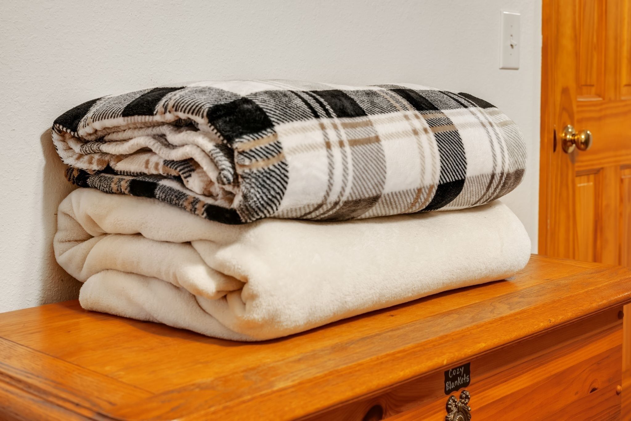 A stack of folded blankets on a wooden table in a room with a wooden door and a light switch on the wall.