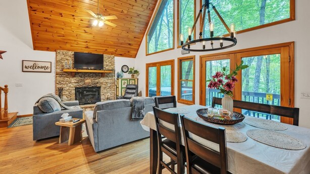 Ein geräumiges Wohnzimmer mit Kamin, eine Essecke mit Tisch für vier Personen und große Fenster mit Blick auf den Wald.